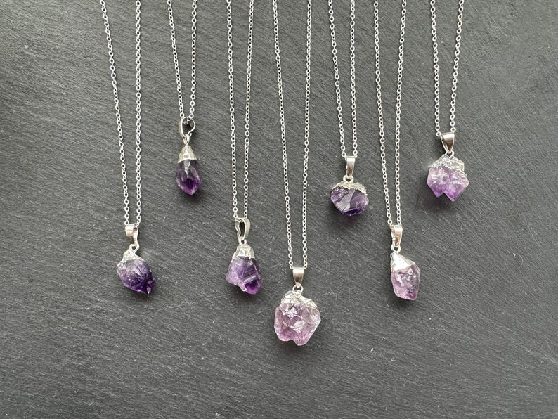 May include: Seven silver necklaces with amethyst crystal pendants. The crystals are all different shapes and sizes, and they have a purple and pink hue.