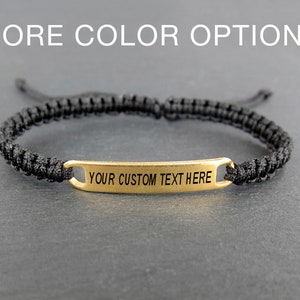 May include: Black braided bracelet featuring a gold-coloured rectangular plate. The plate is engraved with the text "YOUR CUSTOM TEXT HERE". The background includes the text "MORE COLOR OPTIONS".