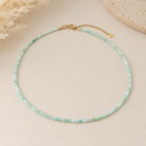 May include: A delicate necklace featuring small, faceted light green beads strung together. The piece includes a gold-toned metal lobster claw clasp and a short adjustable chain extension with a small circular charm at the end of the chain.