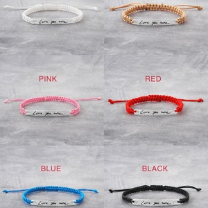 May include: A set of ten bracelets with different coloured cords and a silver bar with the handwritten text "Love you more". The bracelets are displayed on a white background with the colour of each bracelet labelled above it. The colours are white, beige, pink, red, blue, and black.