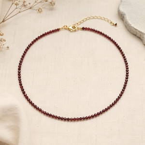 May include: A delicate necklace featuring a single strand of small, faceted, deep red garnet beads. The necklace is finished with a gold-toned metal lobster claw clasp and a short extender chain, creating a circular shape on a neutral fabric.