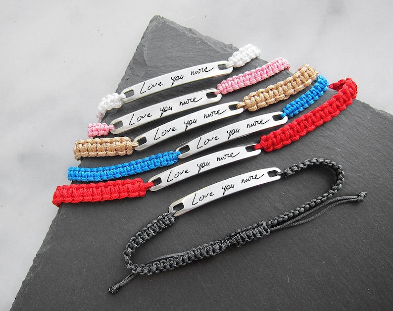May include: Set of seven friendship bracelets with adjustable black cord closures. Each bracelet features a silver metal tag with the handwritten message "Love you more". The bracelets are in various colours, including white, pink, gold, blue, and red.