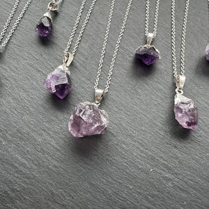 May include: A set of seven silver necklaces with amethyst crystal pendants. The amethyst crystals are all different shapes and sizes, and they have a purple hue.