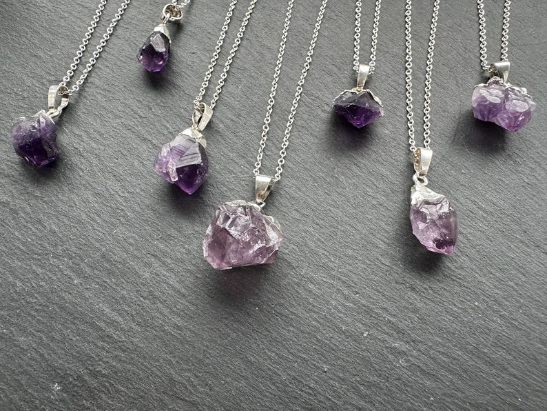 May include: A set of seven silver necklaces with amethyst crystal pendants. The amethyst crystals are all different shapes and sizes, and they have a purple hue.