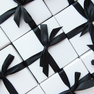 May include: An overhead shot of several white square gift boxes, each tied with a black satin ribbon. The boxes are arranged in a grid pattern, with the black ribbons creating diagonal lines across the image. The overall aesthetic is clean and elegant.