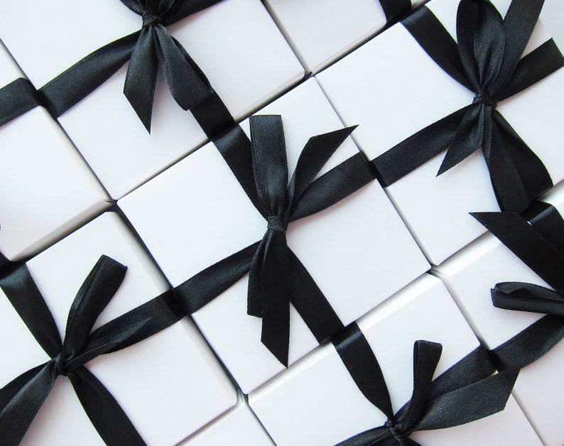 May include: An overhead shot of several white square gift boxes, each tied with a black satin ribbon. The boxes are arranged in a grid pattern, with the black ribbons creating diagonal lines across the image. The overall aesthetic is clean and elegant.