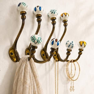May include: A wall-mounted coat rack featuring five curved antique brass metal hooks, each topped with a decorative white ceramic knob painted with colourful floral and leaf patterns. A beige scarf and gold jewellery hang from the hooks.