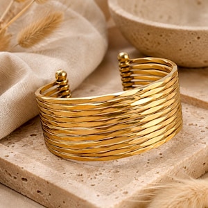 May include: A gold-toned cuff bracelet featuring multiple stacked, hammered-texture bands that connect at the ends with small spherical finials. The design consists of several parallel, thin metal wires that create a wide, layered appearance.