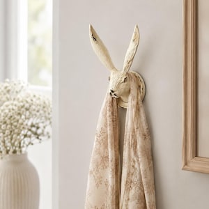 May include: A wall-mounted hook shaped like a rabbit head with long, upright ears. The hook has a distressed, off-white finish and is holding a light-coloured scarf with a brown floral pattern and a thin gold-toned necklace with a small pendant.