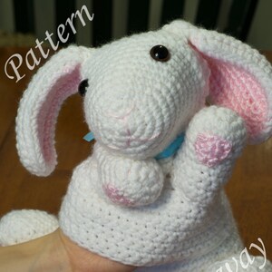 Può includere: Coniglio bianco all'uncinetto con orecchie e piedi rosa. Il coniglio sta alzando una zampa. Il coniglio è seduto su una superficie di legno. Il testo "Pattern" è nell'angolo in alto a sinistra dell'immagine. Il testo "Crochet Gateway Photography" è nell'angolo in basso a destra dell'immagine.