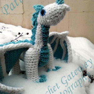 May include: A white and light blue crocheted dragon toy with a blue eye. The dragon is sitting in the snow.