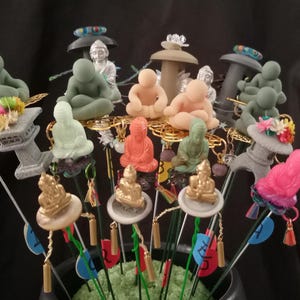 May include: A collection of decorative zen garden figurines, including seated Buddhas and miniature stone lanterns, in various colors. Some figurines are adorned with colorful floral accents and are mounted on wire stems.