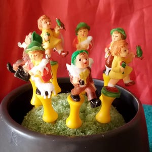 May include: Seven small figurines of children dressed in green and yellow clothing, holding sheep and birds. The figurines are standing on yellow bases and are arranged in a circle on a green surface.