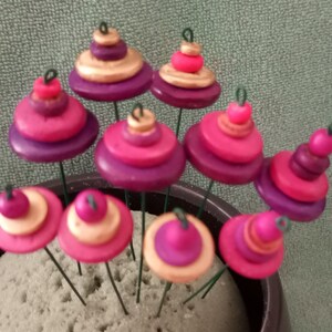 May include: A collection of ten pink, purple, and gold stacked buttons on wire stems. The buttons are arranged in a circular pattern and are displayed in a small pot.