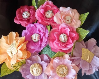 May include: A collection of fabric flowers in various shades of pink, orange, and purple. Each flower features a face-shaped embellishment at its center. Green leaves are interspersed among the blooms, creating a whimsical floral arrangement.