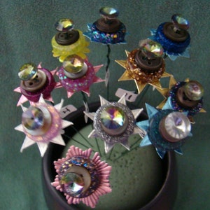 May include: A bouquet of colorful button flowers with sparkly accents. Each flower is made from a button with a different design and color, including blue, green, pink, and gold. The flowers are arranged in a vase with a green background.