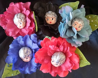 May include: A collection of colorful fabric flower brooches, each featuring a vintage doll face center. The flowers are in shades of pink, black, blue, red, and teal, with green leaves.