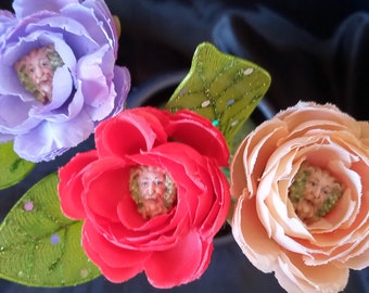 May include: Close-up of three artificial flowers in shades of purple, red, and peach. Each flower has a face in the center. Green leaves with white polka dots are visible. The background is a dark, solid color.