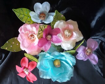 May include: A bouquet of fabric flowers with doll faces in the center of each bloom. The flowers are pink, blue, purple, and white. The flowers are arranged in a circle with green leaves.