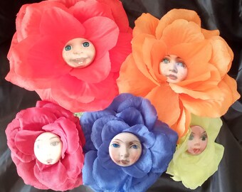 May include: A collection of five fabric flower brooches in vibrant colors: red, orange, pink, blue, and yellow-green. Each flower features a ceramic face at its center, with painted eyes and a smiling expression.