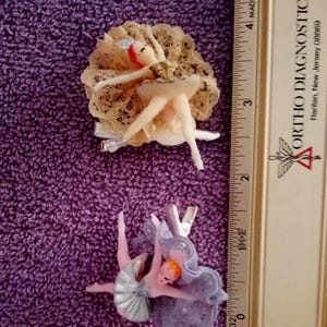 May include: Two vintage ballerina hair clips. One clip features a ballerina with a cream-colored tutu and a lace backdrop. The other clip shows a ballerina in a lavender tutu and a matching lace backdrop. Both clips have silver-colored metal clips.