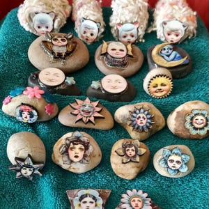 May include: A collection of decorative stones featuring various designs. Some stones have sheep-like figures, while others display faces within floral or celestial motifs. The color palette includes beige, blue, pink, and yellow.