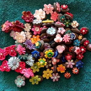 May include: A collection of colorful, flower-shaped embellishments in various sizes and hues, including pink, red, yellow, and blue. Each flower is attached to a small, dark-colored base. The items are arranged on a teal-colored surface.