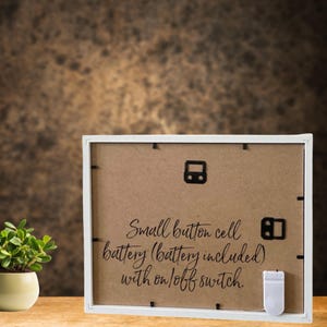 May include: White picture frame with a brown cardboard backing. The back of the frame has text that reads "Small button cell battery (battery included) with on/off switch."