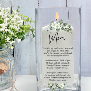May include: A white candle with a floral design and the word "Mom" on it. The candle is in a clear glass jar. The text on the candle reads: "You held my hand when I was small You caught me when I fell You're the hero of my childhood And my later years as well And every time I think of you My heart still fills with pride Though I'll always miss you I know you're by my side In laughter and in sorrow In sunshine and through rain I know you're watching over me Until we meet again"