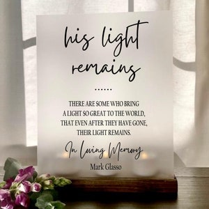 May include: A white sign with black text that reads "his light remains" followed by a quote about the lasting impact of a person's light. The quote reads "There are some who bring a light so great to the world, that even after they have gone, their light remains." The sign also reads "In Loving Memory Mark Glasso".