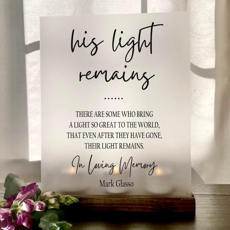 May include: A white sign with black text that reads "his light remains" followed by a quote about the lasting impact of a person's light. The quote reads "There are some who bring a light so great to the world, that even after they have gone, their light remains." The sign also reads "In Loving Memory Mark Glasso".