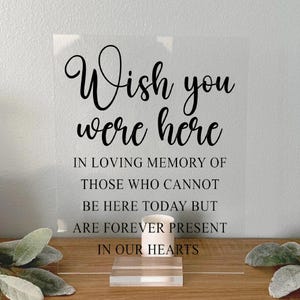 May include: A clear acrylic sign with the text "Wish you were here" in black cursive font. The sign also includes the text "In loving memory of those who cannot be here today but are forever present in our hearts."