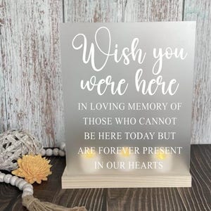 May include: A frosted glass sign with the text "Wish you were here" in white lettering. The sign also includes the text "In loving memory of those who cannot be here today but are forever present in our hearts".
