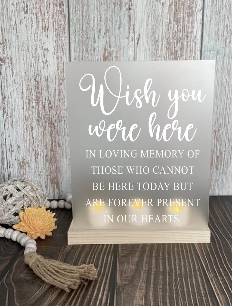 May include: A frosted glass sign with the text "Wish you were here" in white lettering. The sign also includes the text "In loving memory of those who cannot be here today but are forever present in our hearts".