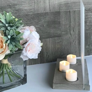 May include: A gray wooden candle holder with three white pillar candles. The candle holder is in front of a clear acrylic panel and a bouquet of pink and green flowers in a glass vase.