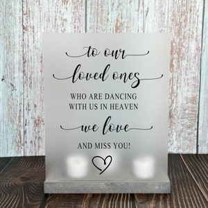 May include: A white sign with black text that reads "to our loved ones who are dancing with us in heaven we love and miss you!" with a heart symbol at the bottom. The sign is sitting on a wooden base.