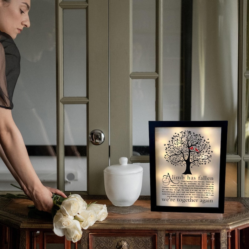May include: A black framed light-up sign with a silhouette of a tree with red birds and the text "A limb has fallen, we're together again".