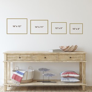 Op de afbeelding: Vier ingelijste foto's aan een witte muur, met de afmetingen van elke foto eronder. De foto's zijn 16 inch bij 12 inch, 14 inch bij 11 inch, 12 inch bij 9 inch en 10 inch bij 8 inch. Voor de muur staat een houten consoletafel met twee laden en een onderplank. Op de tafel staan een schelpenkom, een stapel boeken, een geweven mand en een decoratief metalen beeld.