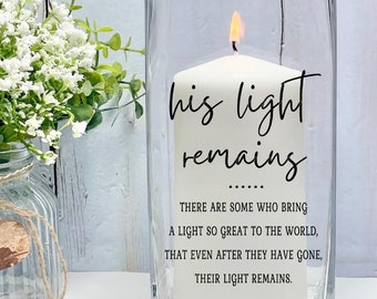 Puede incluir: Una vela blanca encendida dentro de un jarrón de vidrio transparente. La vela tiene las palabras "his light remains" impresas, junto con una cita. Un pequeño ramo de flores blancas se encuentra a la izquierda del jarrón.