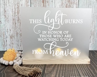 May include: A white frosted glass sign with the text "THIS light BURNS IN HONOR OF THOSE WHO ARE WATCHING TODAY FROM heaven" with three small lights behind the text. The sign is on a wooden base.
