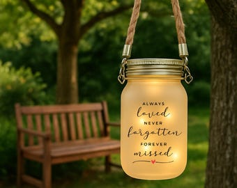 May include: A frosted glass jar lantern with a gold lid and a rope handle, hanging from a tree branch. The jar has the words "Always loved, never forgotten, forever missed" in black script, with a small red heart.