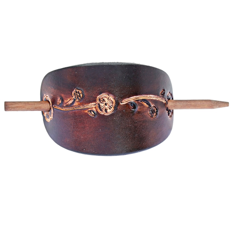 May include: A brown leather hair stick with a floral design and a wooden dowel. The stick is curved and has a decorative floral pattern.