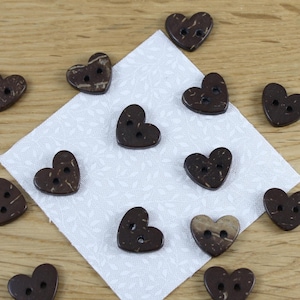 May include: Heart-shaped wooden buttons in dark brown, scattered on a patterned white fabric and a wooden surface. Each button has two holes for sewing. The buttons are made from wood.