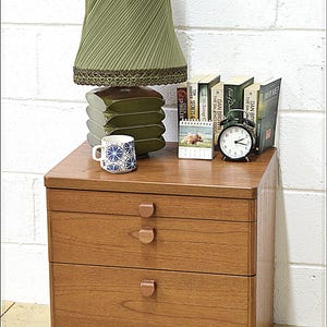 May include: A wooden bedside cabinet with three drawers, featuring a green lampshade, books, a mug, and a clock. The lamp has a pleated shade and sits above a stack of decorative elements. Books include titles like "The Da Vinci Code".