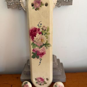May include: Cream-coloured, elongated decorative piece with a floral rose design in pink and white. Two matching round knobs with rose patterns are at the bottom. The piece is mounted on a grey, stepped base.