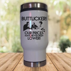 May include: White travel mug with a black graphic of three people sitting at a table. The text on the mug reads "Buttlicker! Our prices have never been lower!"