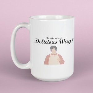 Puede incluir: Taza de cerámica blanca con un asa grande. La taza presenta el texto "In the most Delicious Way!" y una ilustración de una persona con un delantal rosa. La taza está sobre un fondo rosa.