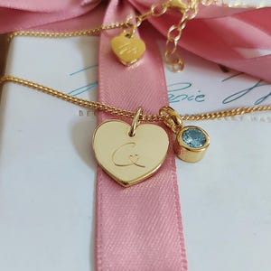 May include: A gold-toned necklace featuring a heart-shaped pendant engraved with the letter C and a small heart, accompanied by a round light blue gemstone charm. The chain includes a small heart-shaped tag near the clasp for adjustment.
