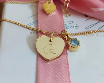 May include: A gold-toned necklace featuring a heart-shaped pendant engraved with the letter C and a small heart, accompanied by a round light blue gemstone charm. The chain includes a small heart-shaped tag near the clasp for adjustment.