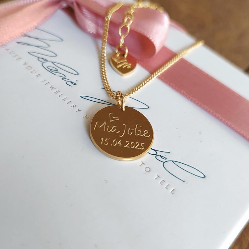 May include: A gold-colored necklace with a round pendant engraved with the name "Mia Jolie" and the date "15.04.2025". The necklace is on a white box with a pink ribbon.
